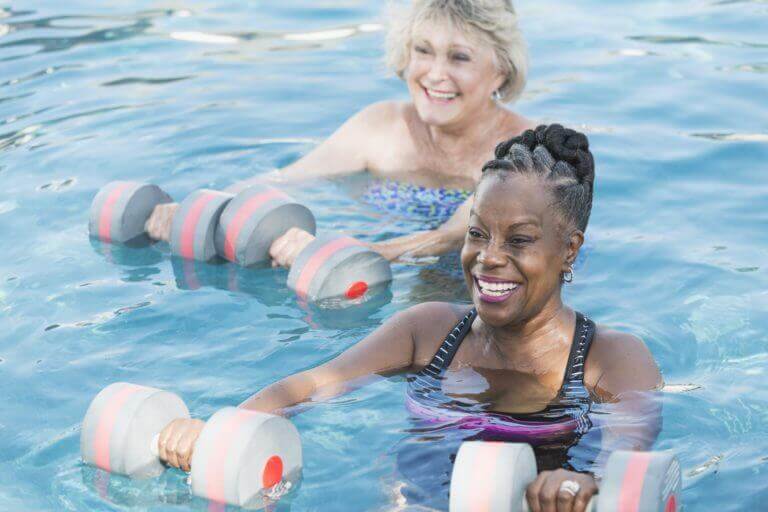 Two senior women smiling and exercising in a pool with foam dumbbells during a water aerobics session.