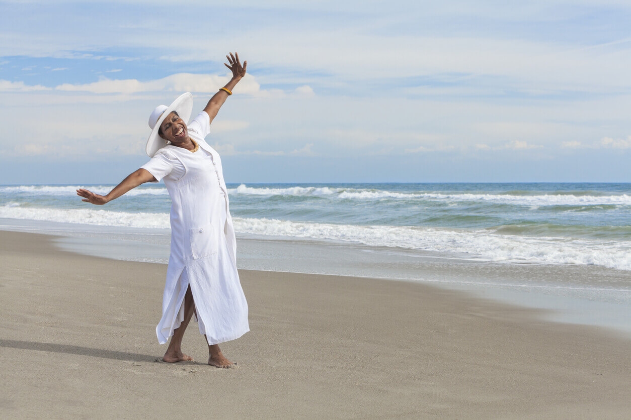 Elderly woman wearing white dress and hat joyfully poses on a sandy beach in front of the ocean.