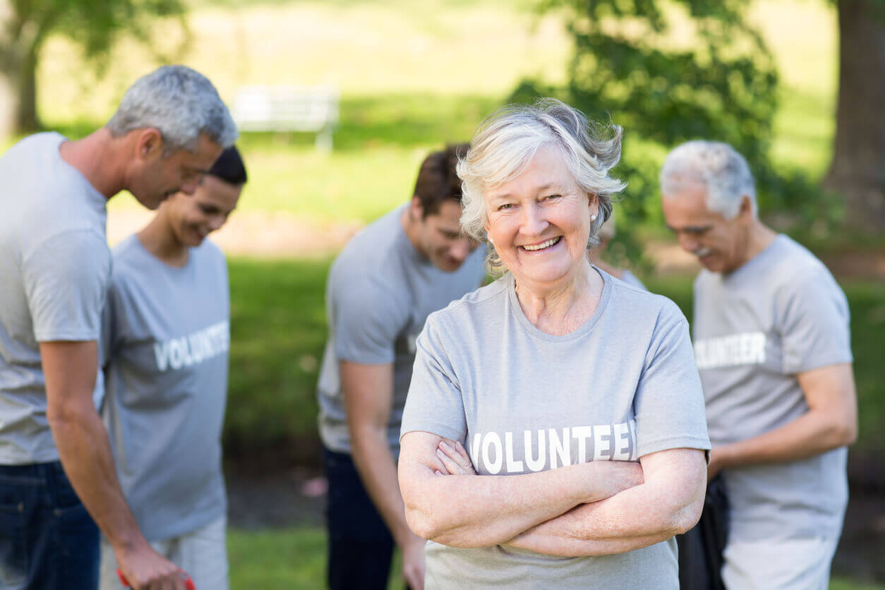 Smiling senior volunteer with crossed arms, surrounded by other volunteers in a park.