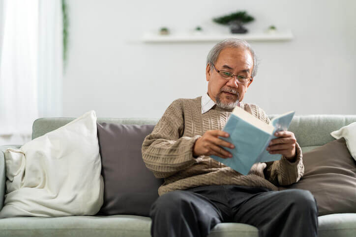 Elderly man reading a book while sitting on a sofa in a cozy living unit.