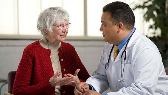 Elderly woman speaking with a doctor in a well-lit room, discussing medical care.