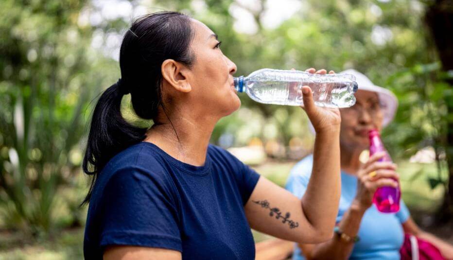 A woman drinks from a water bottle while sitting on a bench outdoors with another person.