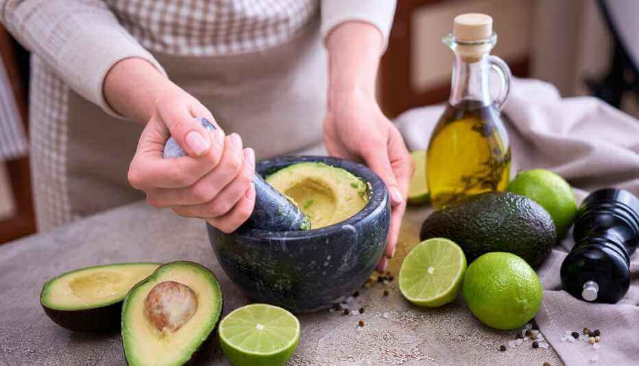 Person mashing avocados in a mortar with limes, olive oil, and pepper on the table.