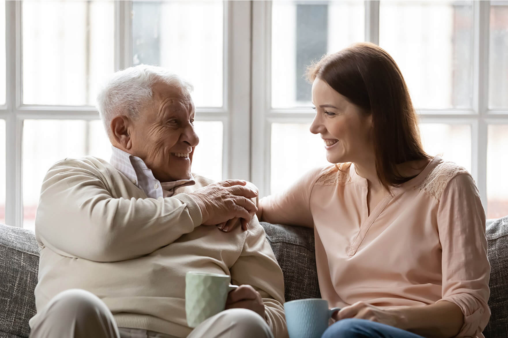 Elderly man and young woman smiling while sitting on a sofa, holding mugs