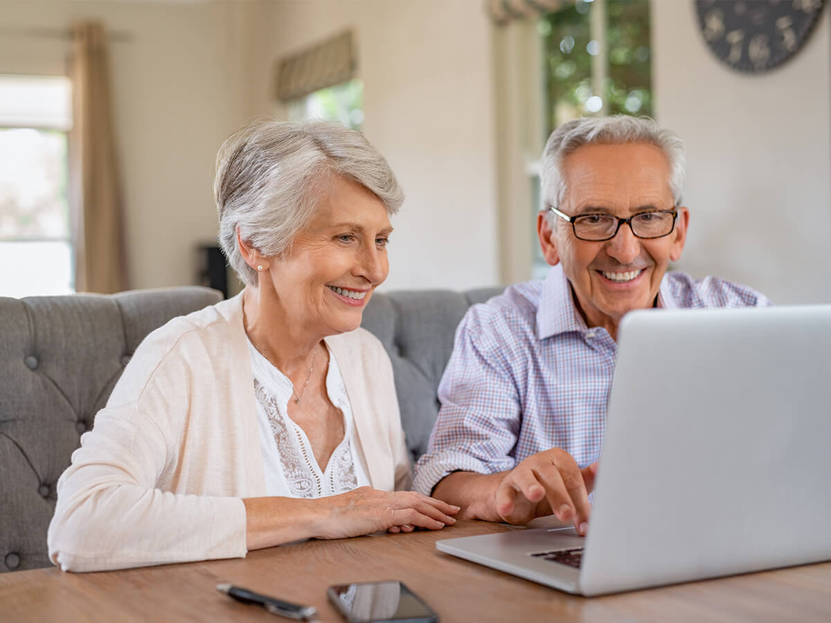 Smiling elderly couple using a laptop at a wooden table in a bright living room.