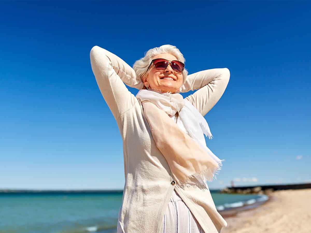 Elderly woman smiling on a sunny beach, wearing sunglasses and a light scarf.