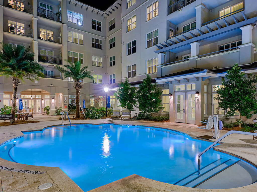 Illuminated swimming pool at night with adjacent units and palm trees in the background.
