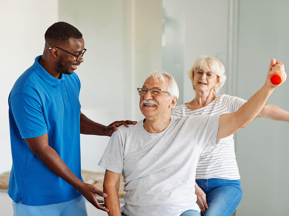 Senior man exercises with a caregiver while a woman guides him at a living community.