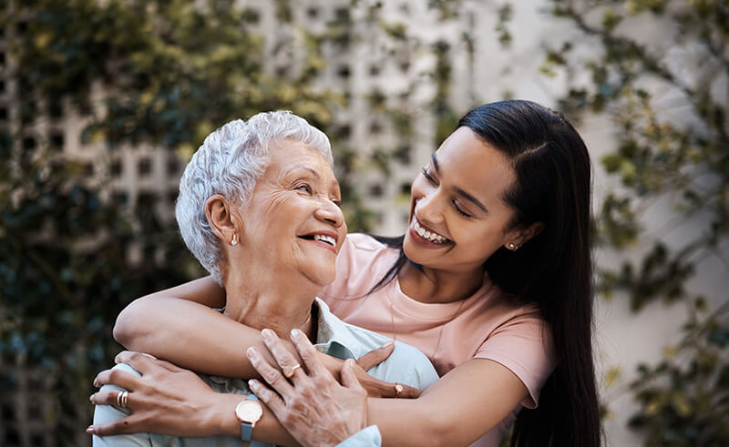 Elderly woman and young woman embrace outdoors, both smiling warmly.
