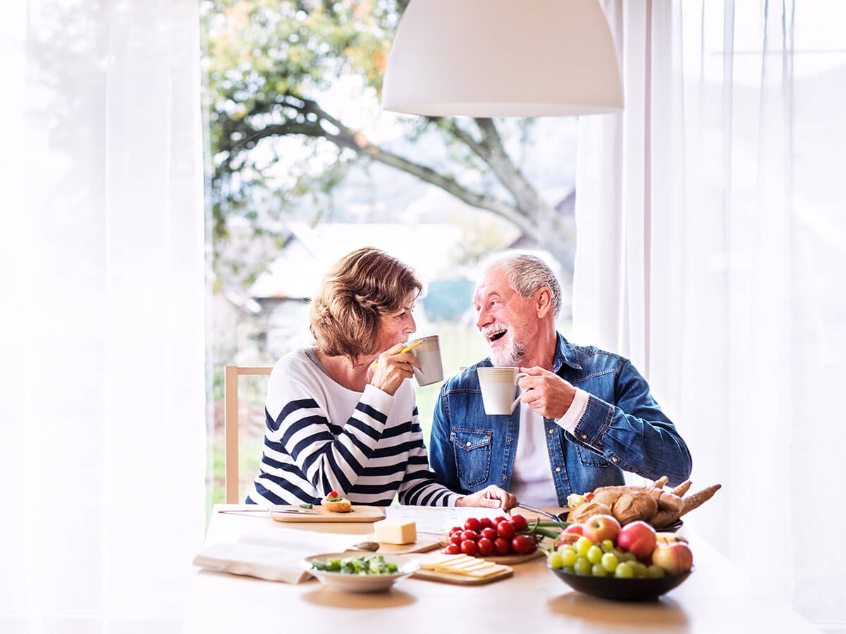 Smiling couple enjoying breakfast with fruits and mugs at a sunny window.