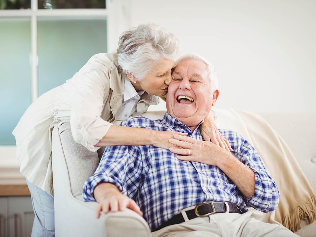 Elderly woman kisses laughing man seated on a sofa in a bright room.