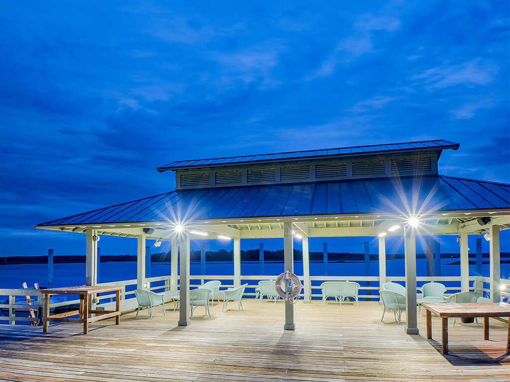 Outdoor pavilion by the water at dusk with seating and bright lighting.