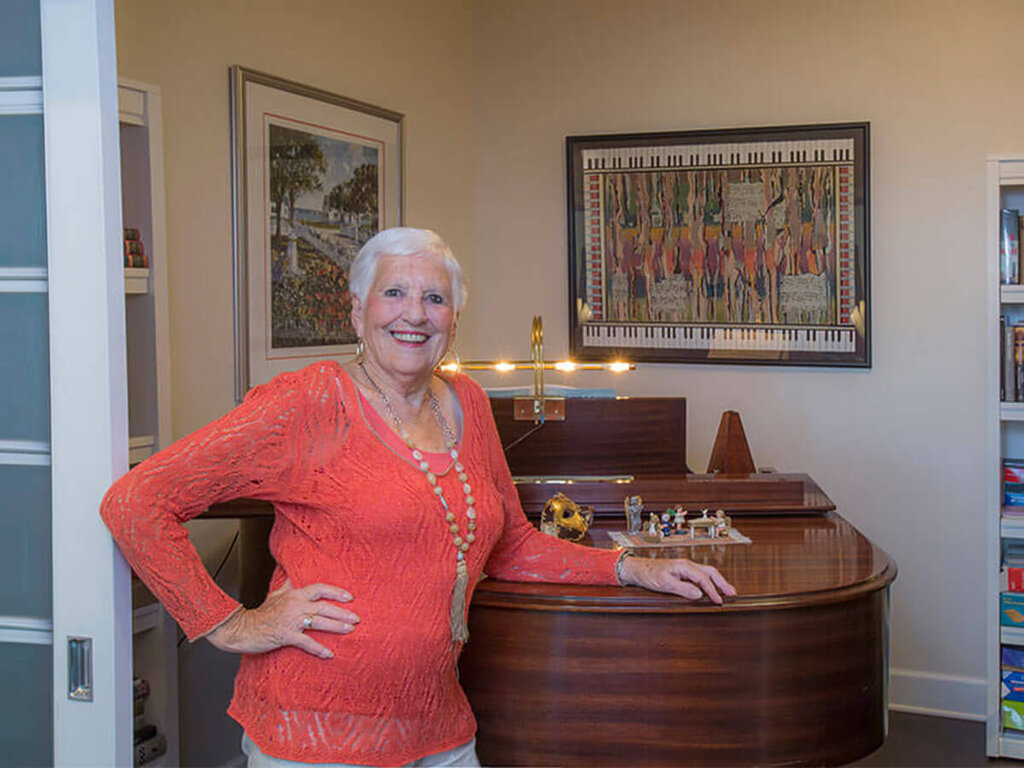 Smiling woman in orange sweater stands by wooden piano in a warmly decorated room.