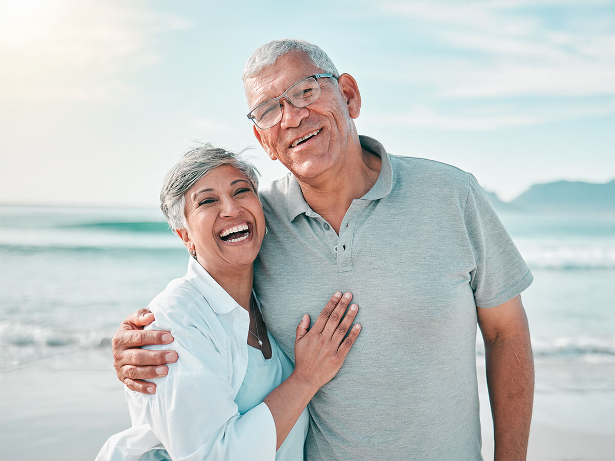 Smiling senior couple embracing on a beach with ocean and sky in the background.