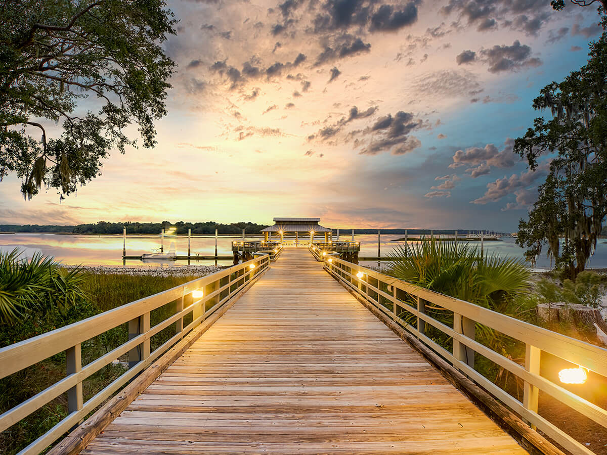 Long wooden pier leading to a serene waterfront at sunset with a colorful sky.